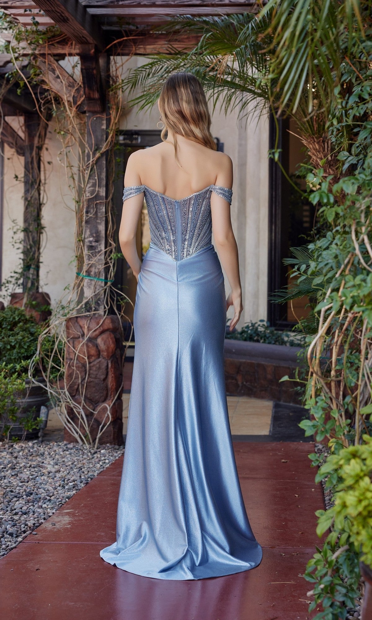 A woman stands outside with her back to the camera, wearing the Nox Anabel F1727 long prom dress in light blue, featuring an off-the-shoulder design and corset bodice, surrounded by greenery and a rustic building.